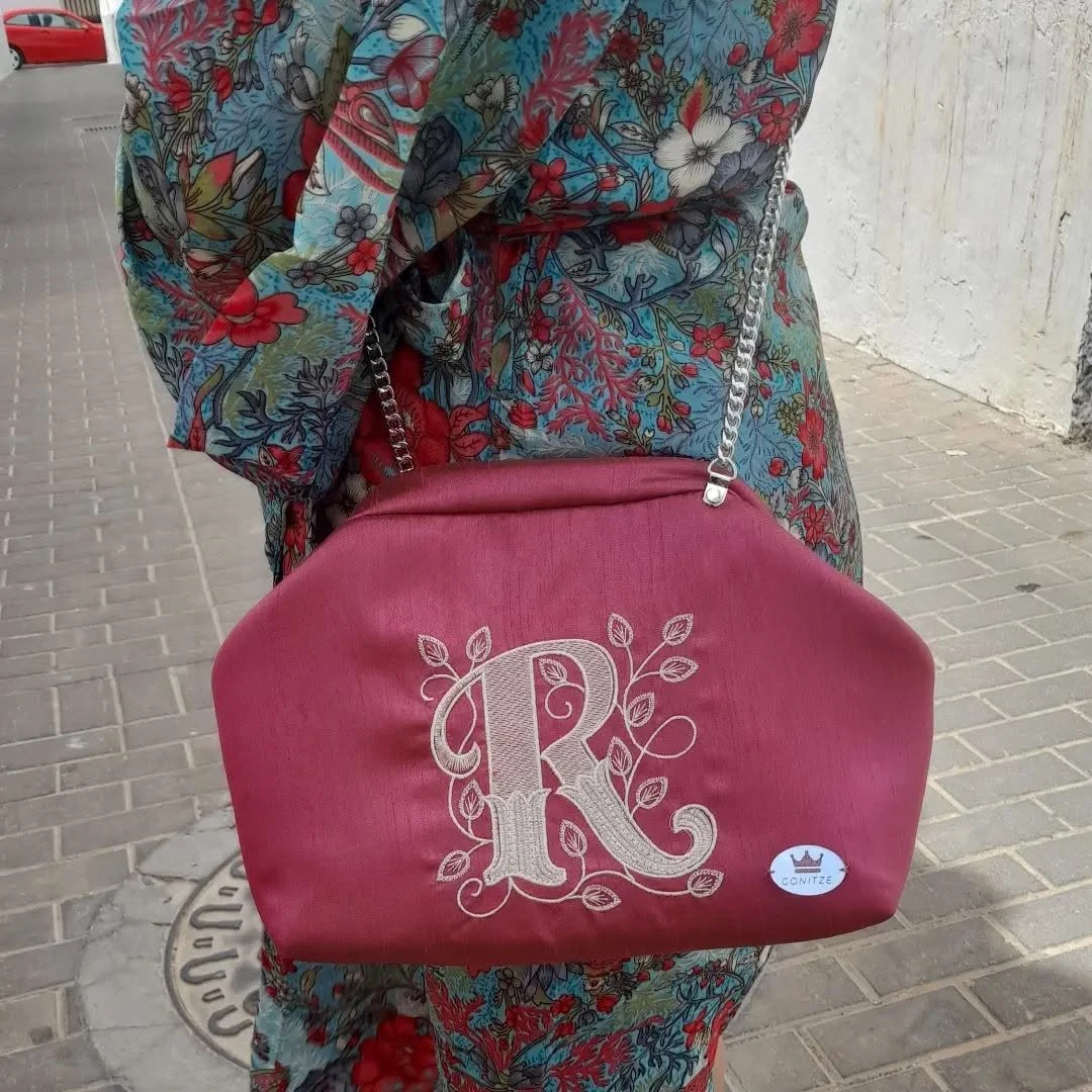 Bolso Gonitze color vino con R bordada, ramas, cadena plateada y sello ovalado, llevado al hombro